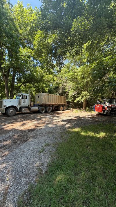 White truck and tan dumpster trailer on a gravel drive under trees, with a red vehicle at right.