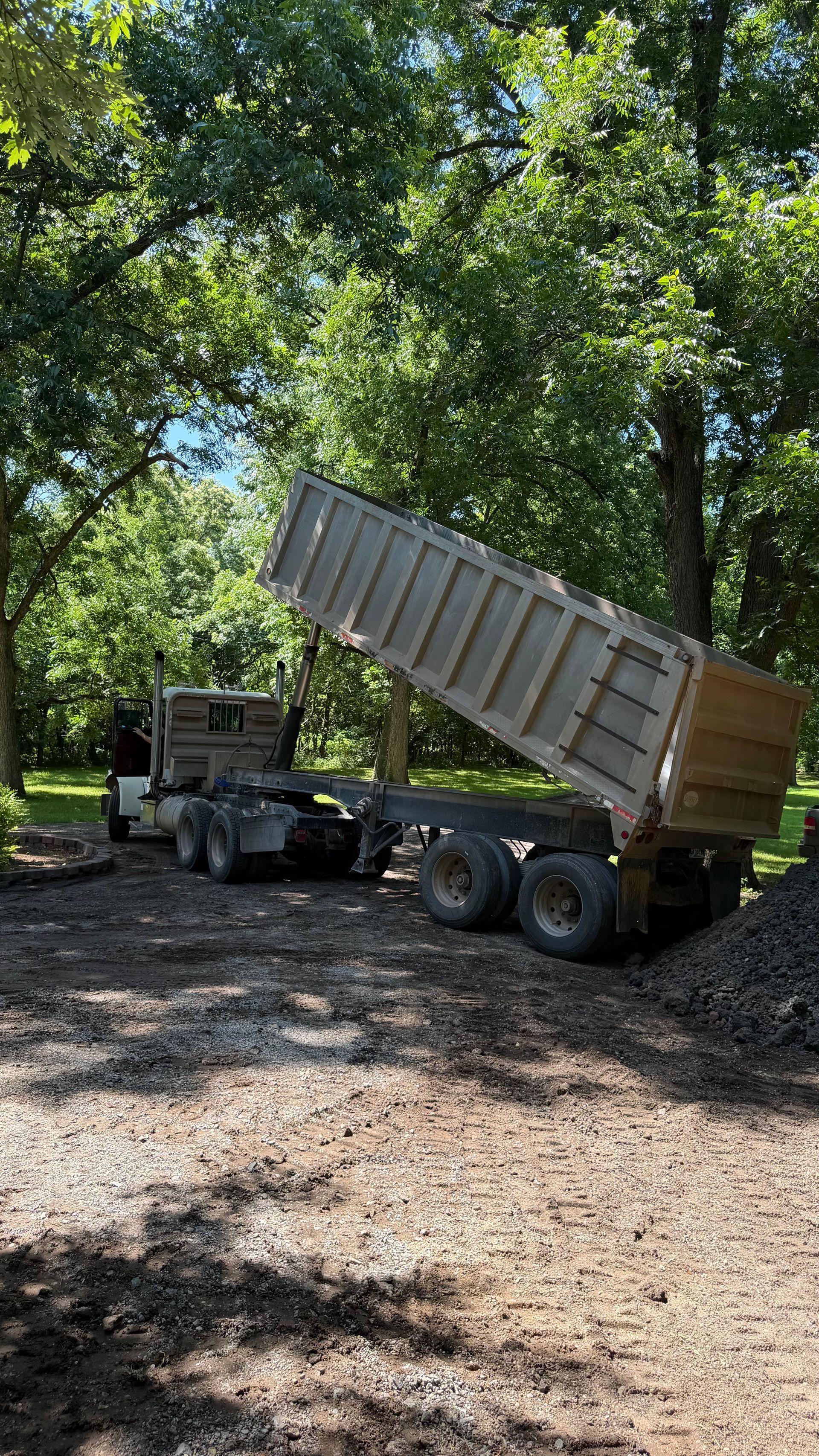Dump truck raising its bed on a gravel driveway under trees