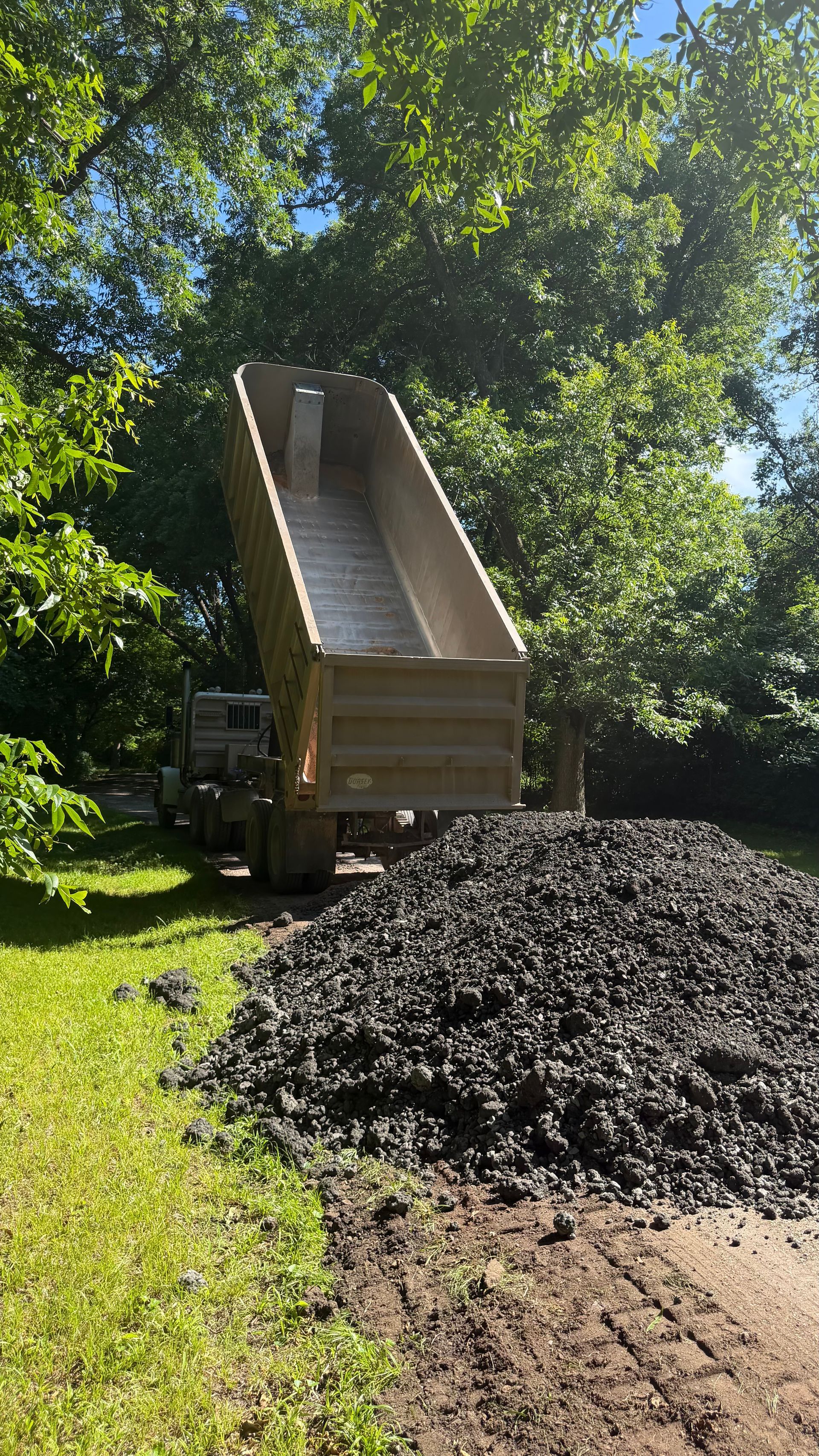 Construction dump truck unloading a large pile of dark gravel in a sunny yard with trees.