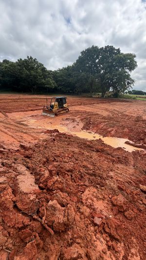 Small excavator working in a muddy red field under cloudy skies, with trees in the background
