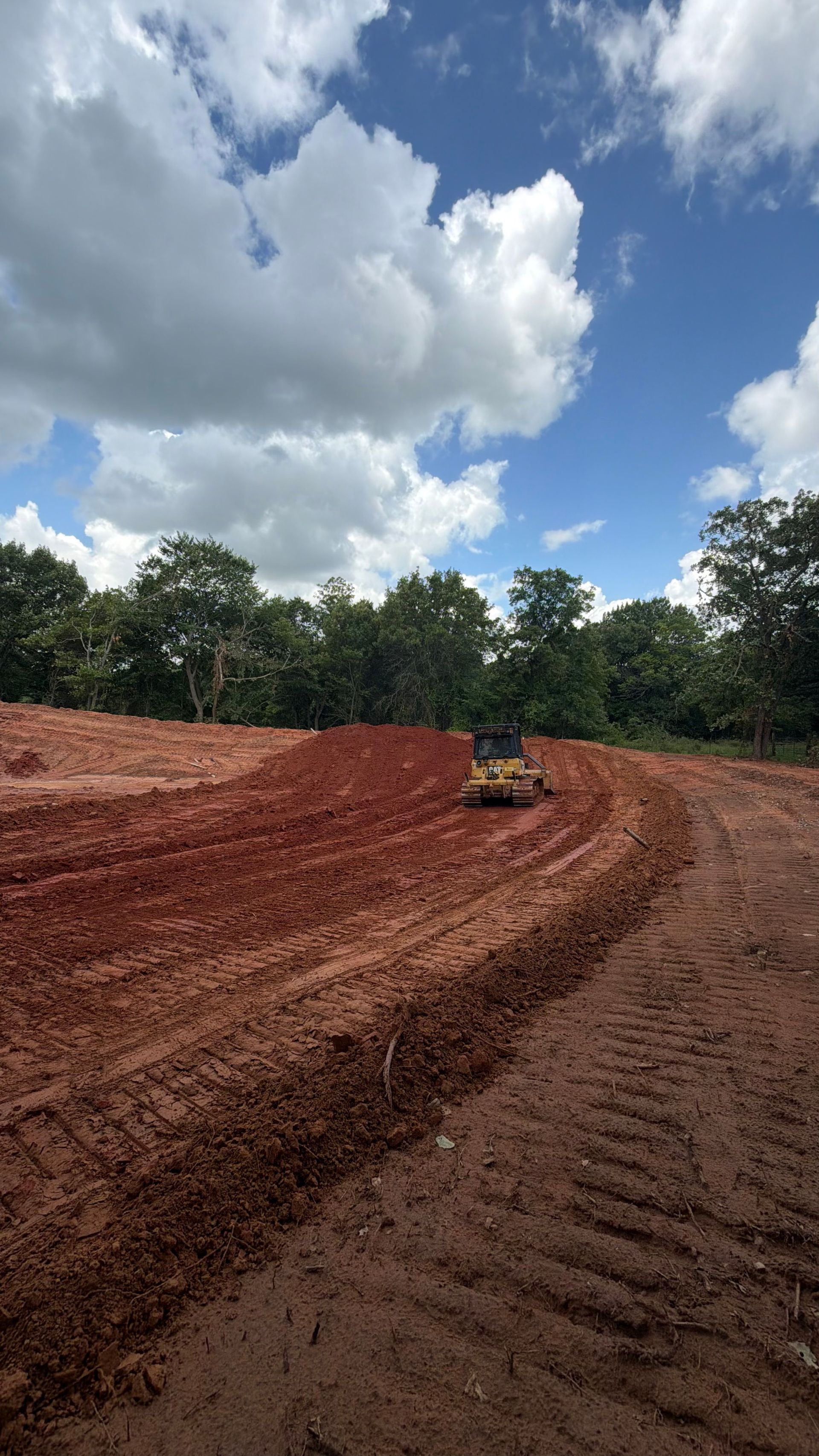 Bulldozer grading a red dirt site under a cloudy sky, with trees along the edge