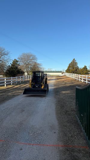 Small excavator on a gravel path between white fences under a clear blue sky.