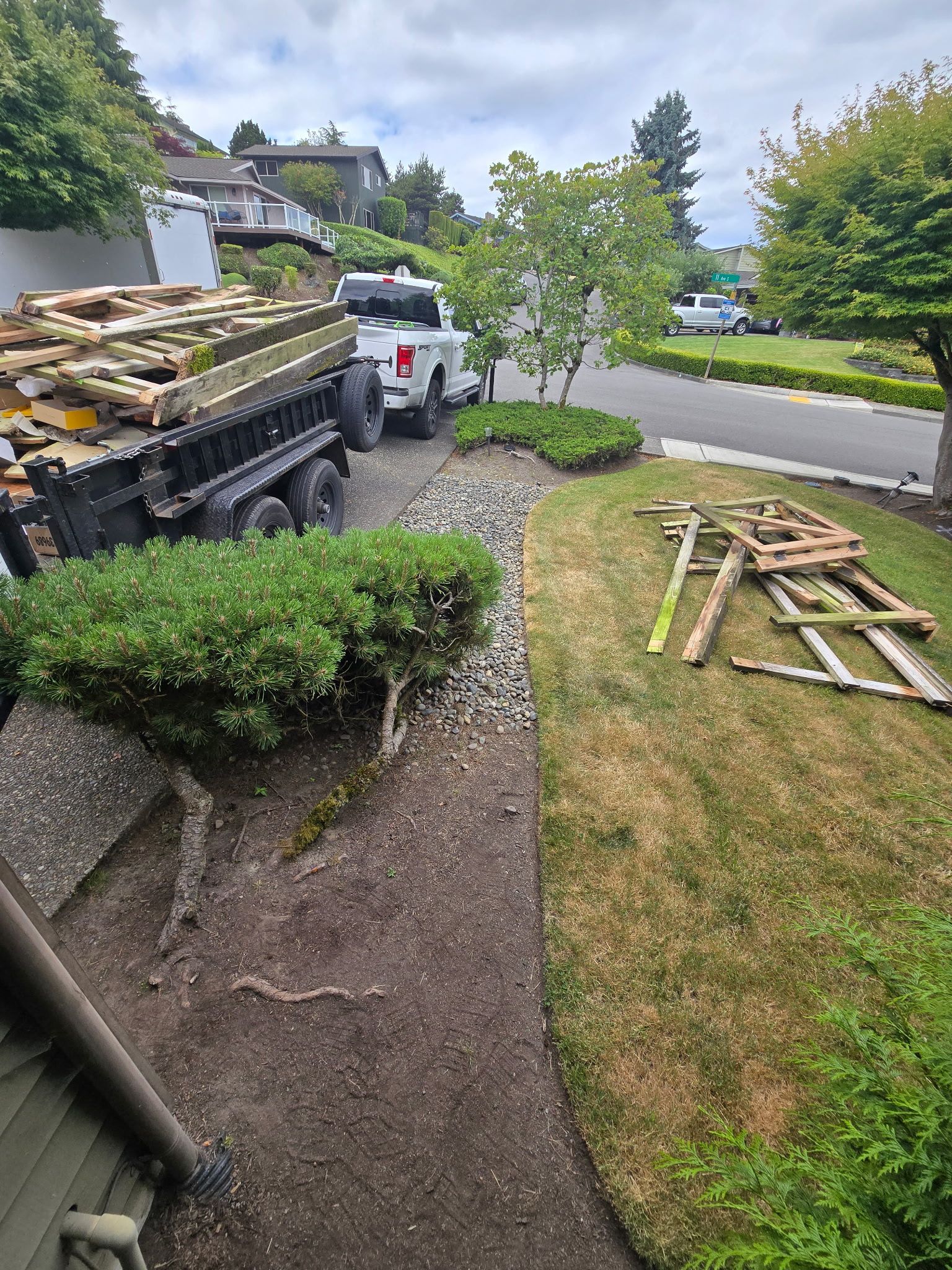 Front yard with white truck, stacked lumber on a trailer, gravel path, trimmed shrubs, and lawn.