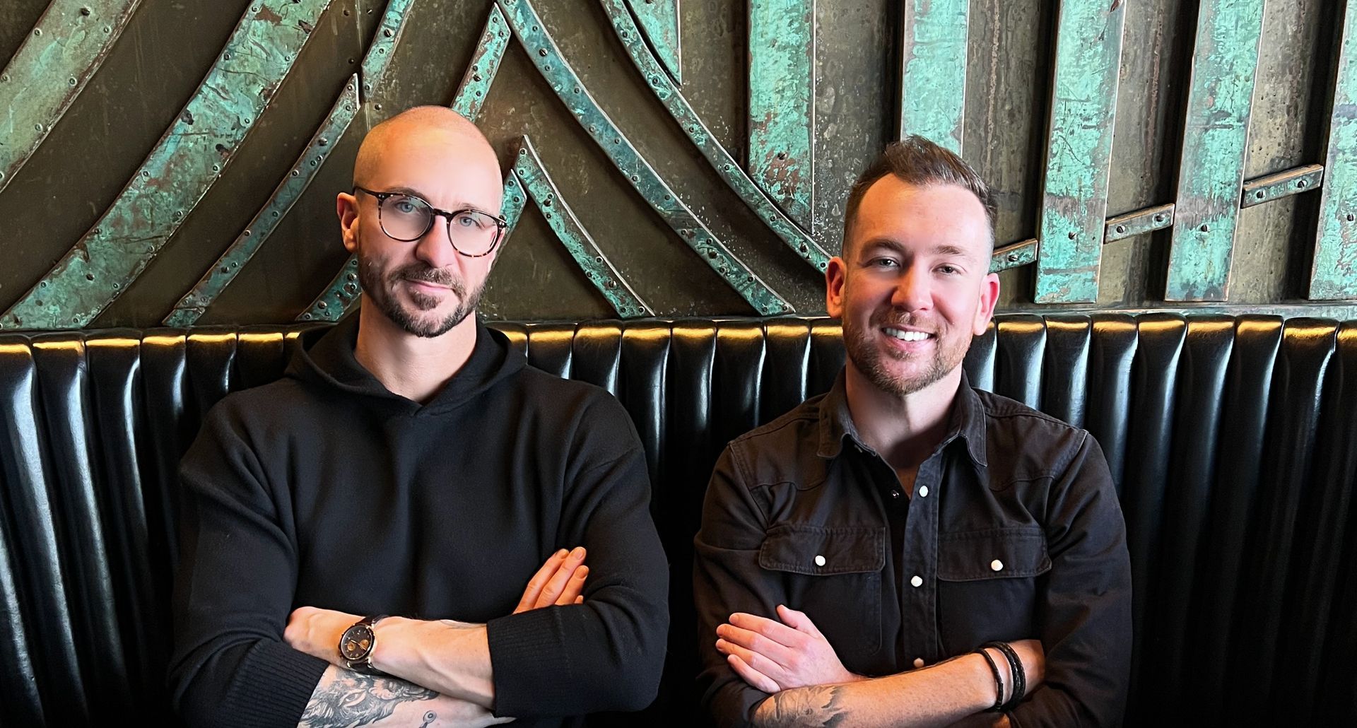 Two men sit side-by-side, arms crossed. One wears glasses and a black sweater; the other a black shirt and bracelets.