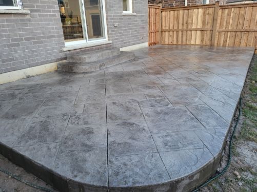 Stamped concrete patio beside a brick house and wooden fence, with a rounded edge.