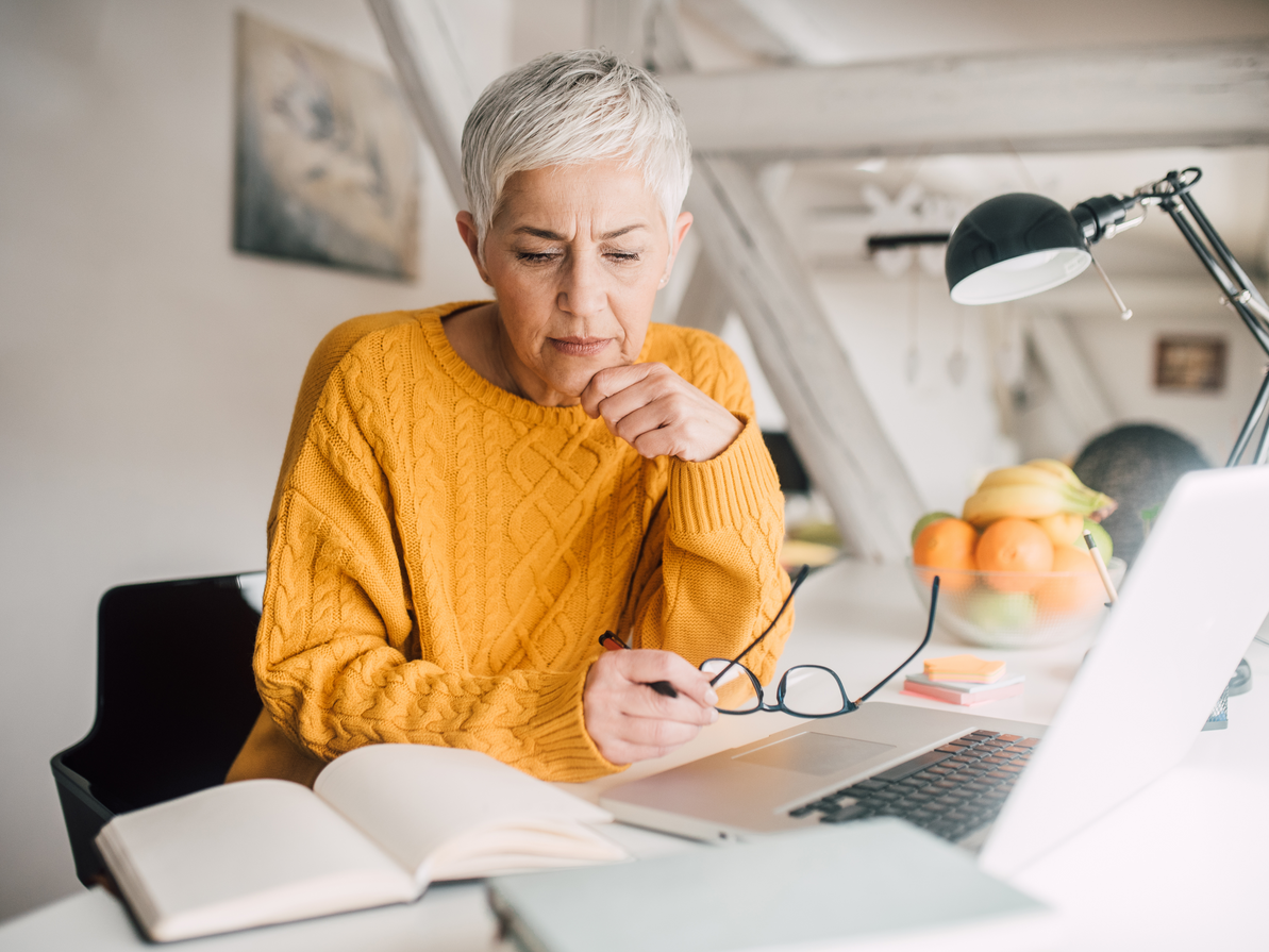 Woman with gray hair in a yellow sweater thoughtfully looks at laptop with glasses in hand; books and fruit nearby.