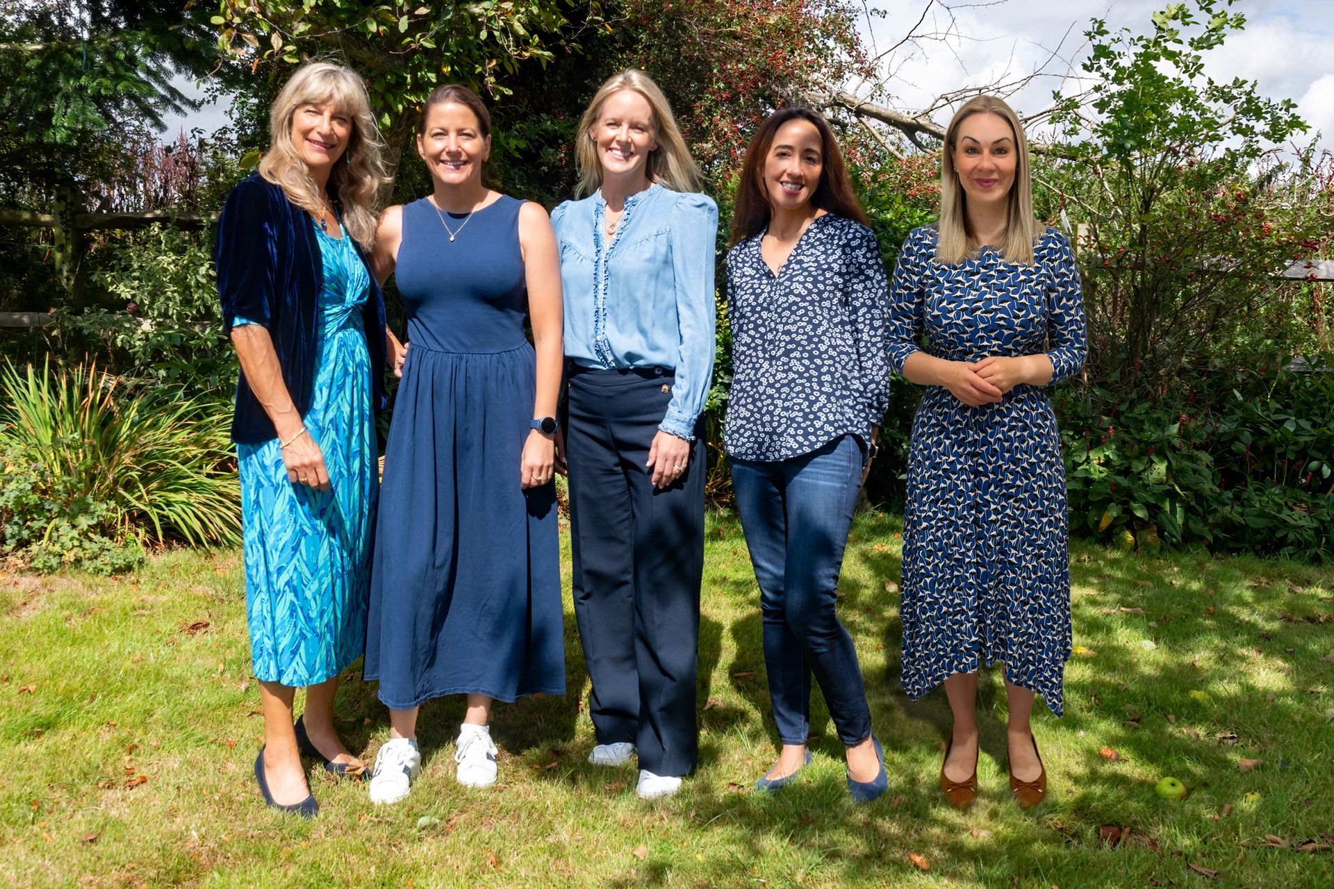 Five women in various shades of blue clothing, standing outside on a grassy lawn.