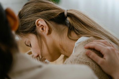 Woman with head down, comforted by a person's hand on her shoulder; beige tones.