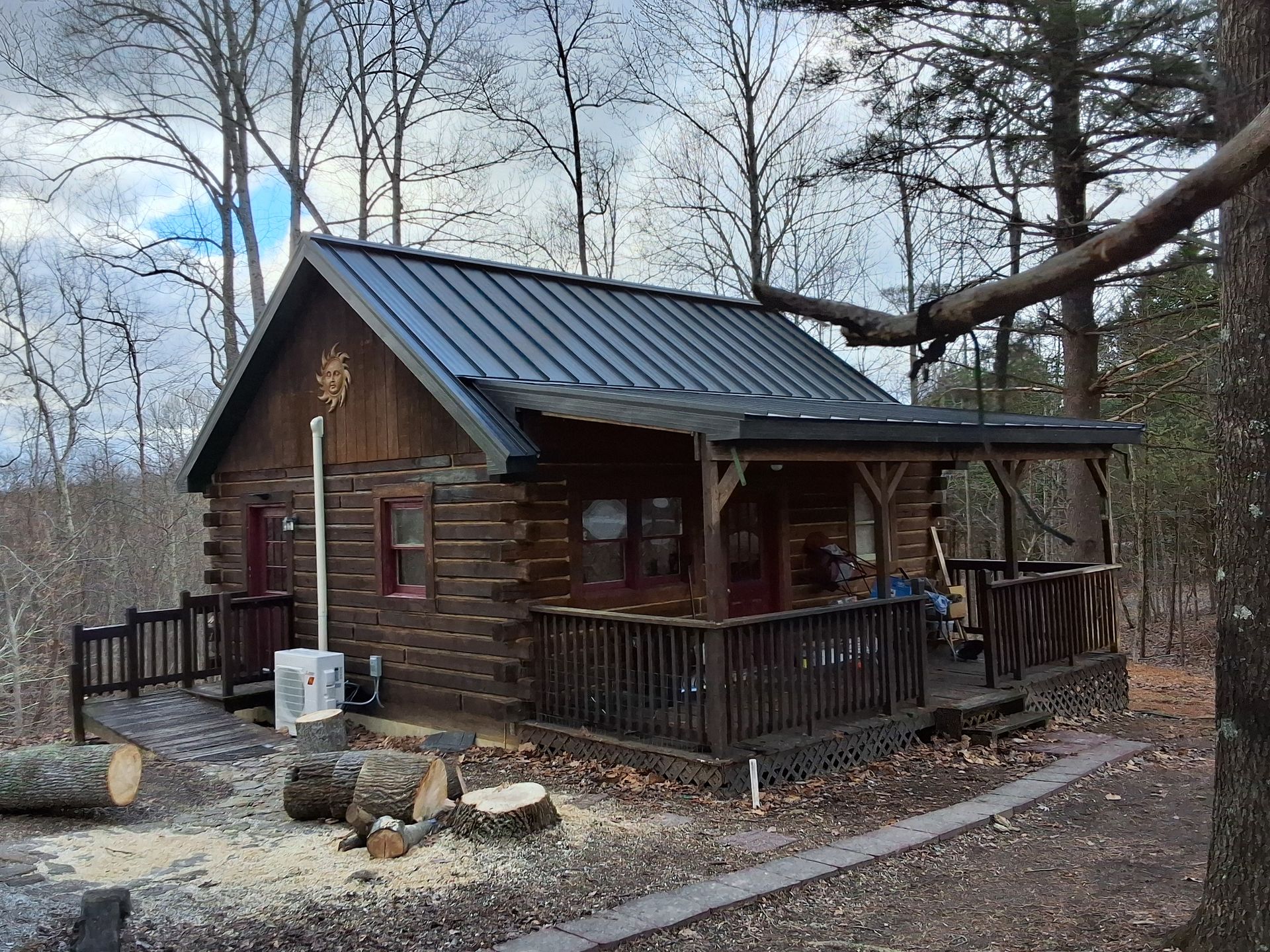 Small rustic cabin with a porch in a wooded area, with bare trees and a gravel path.