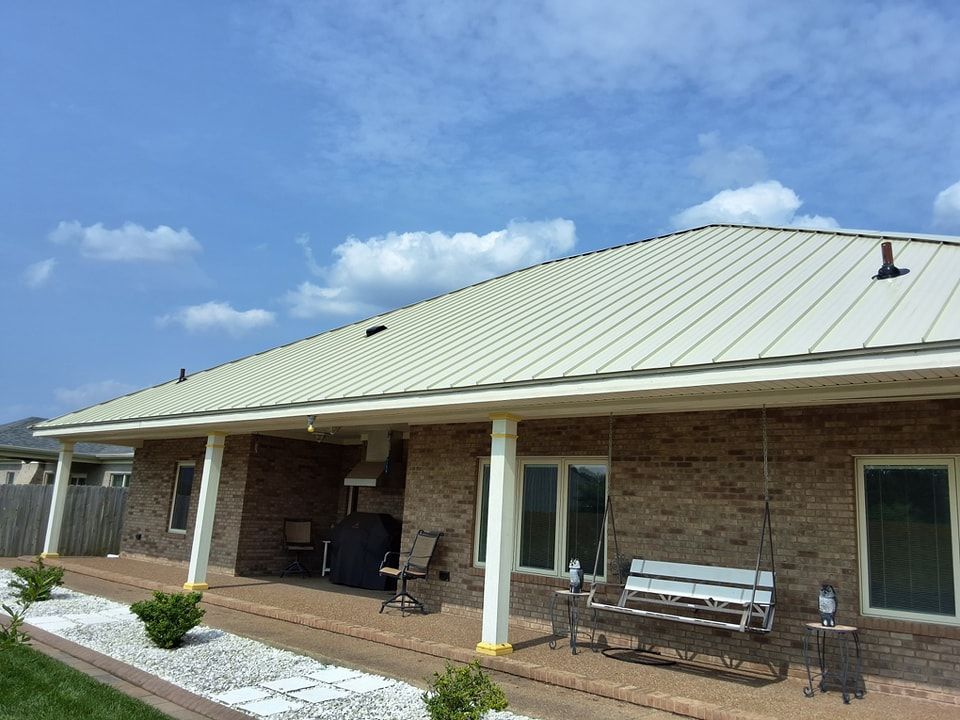 Brick house with a light metal roof, covered porch, and bench under a blue sky.