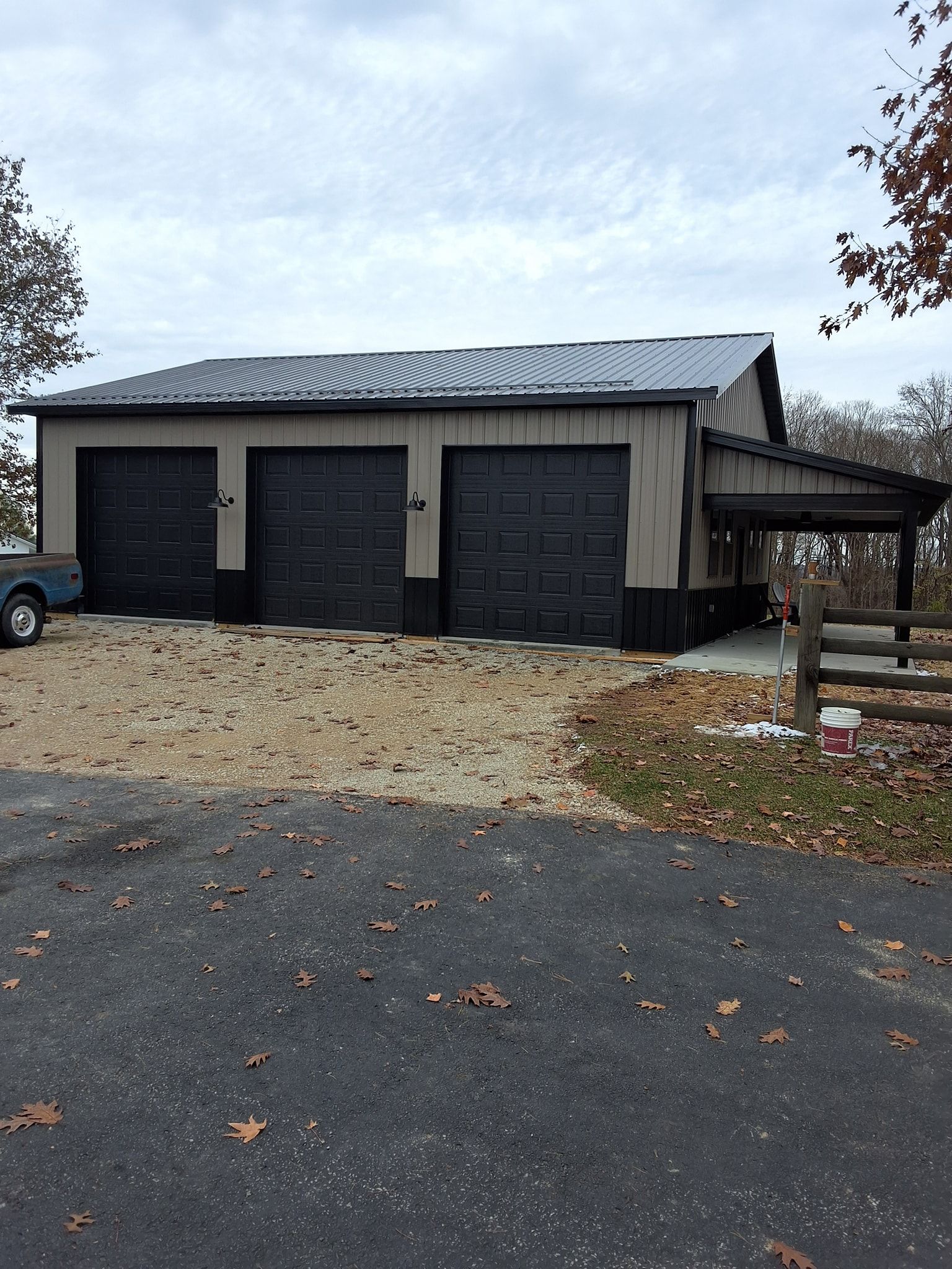 Three black garage doors on a metal workshop with a gravel driveway and cloudy sky