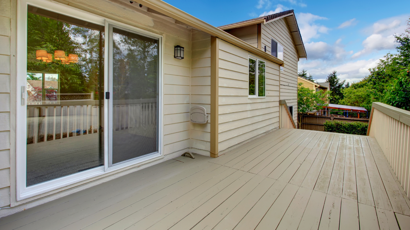 Wide beige deck beside a house, with sliding glass doors and railing overlooking trees and sky