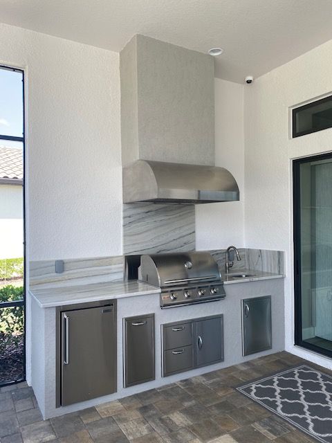 Outdoor kitchen with stainless steel grill, gray cabinets, and hood against white walls and tiled floor