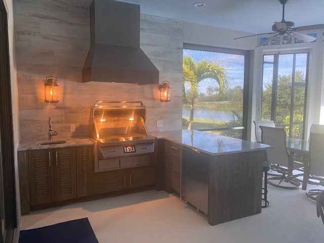 Outdoor kitchen with stainless grill and black countertop beside a pool and palm view