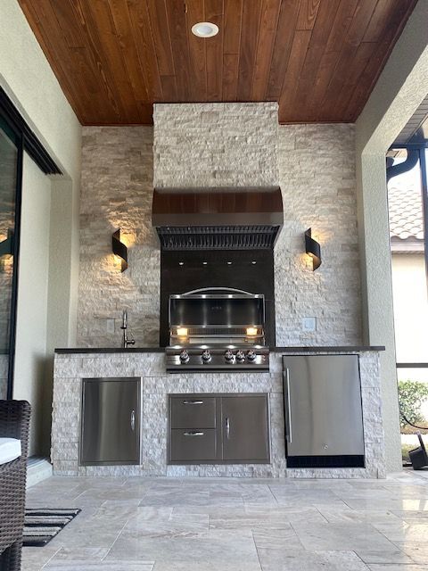 Outdoor kitchen with built-in grill, stone backsplash, dark wood ceiling, and wall sconces