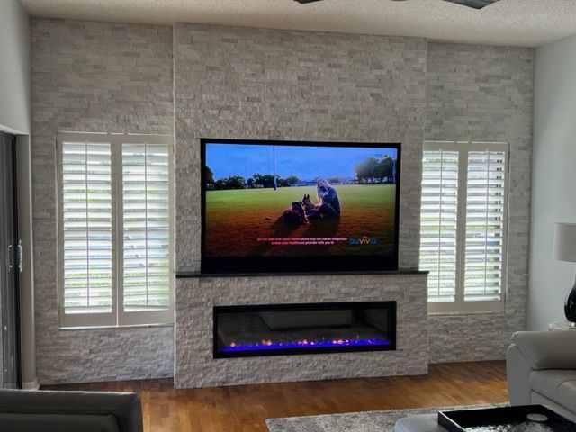 Living room with wall-mounted TV above a linear fireplace, flanked by white shutters and gray stone surround