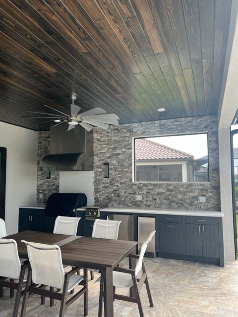 Covered outdoor dining area with wood ceiling, stone wall, table, chairs, and grill