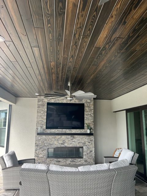 Covered patio with wicker seating around a stone fireplace and TV under a dark wood ceiling