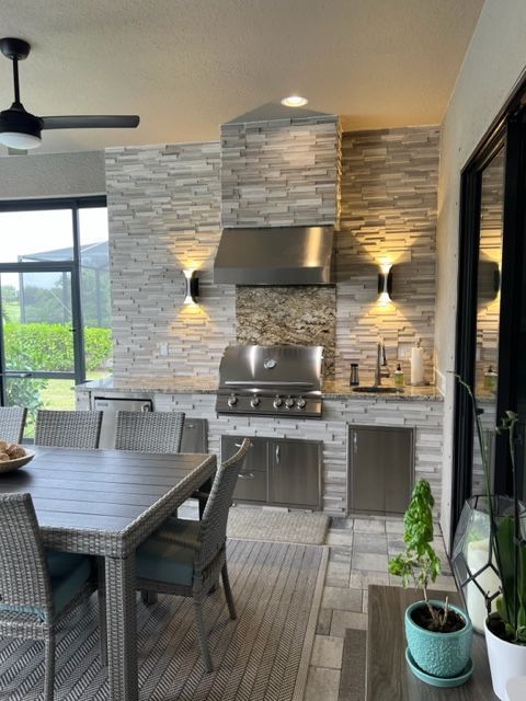 Modern outdoor kitchen with stainless grill, stone backsplash, dining table, and potted plant near large windows