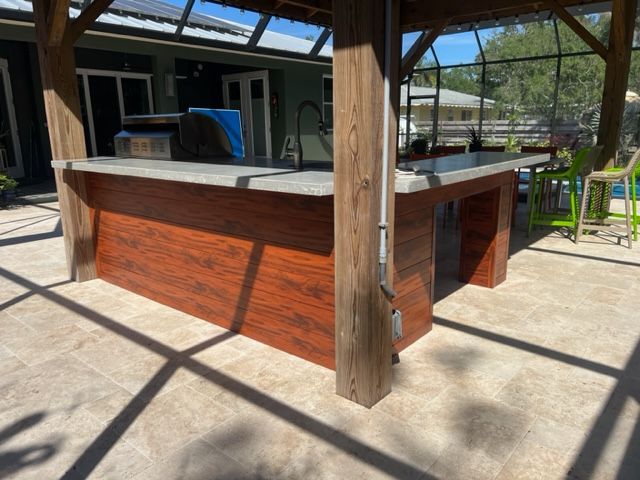Outdoor wooden bar under a shaded patio with a concrete countertop and stools nearby