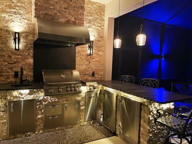 Outdoor kitchen with stainless grill, brick backsplash, and blue-lit patio dining area at night