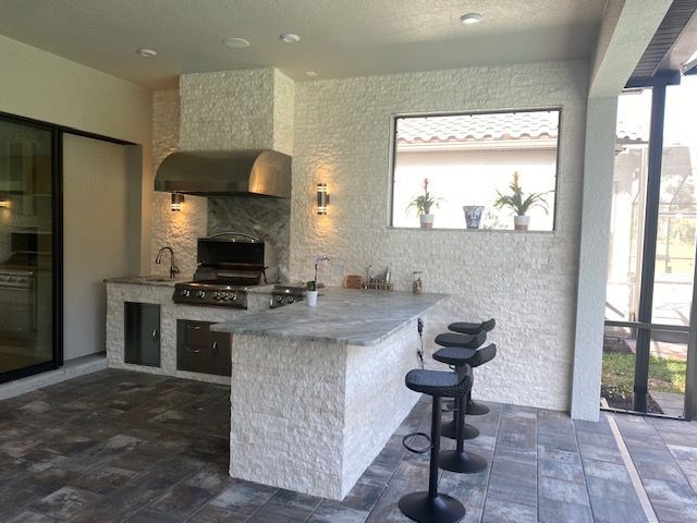 Outdoor kitchen and bar area with built-in grill, stone counters, and three black stools under a covered patio