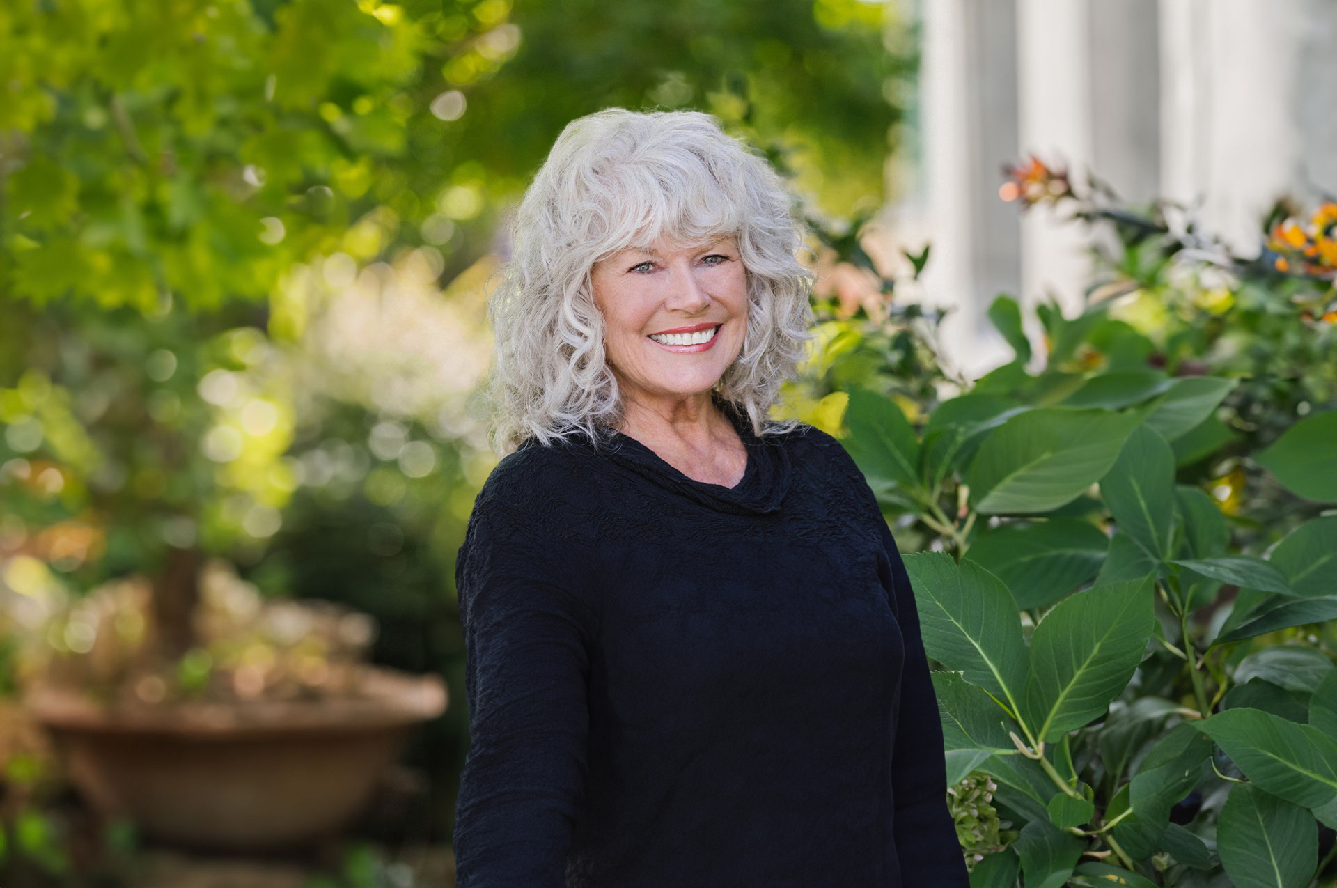 Smiling woman with curly white hair in a black top stands in a leafy garden