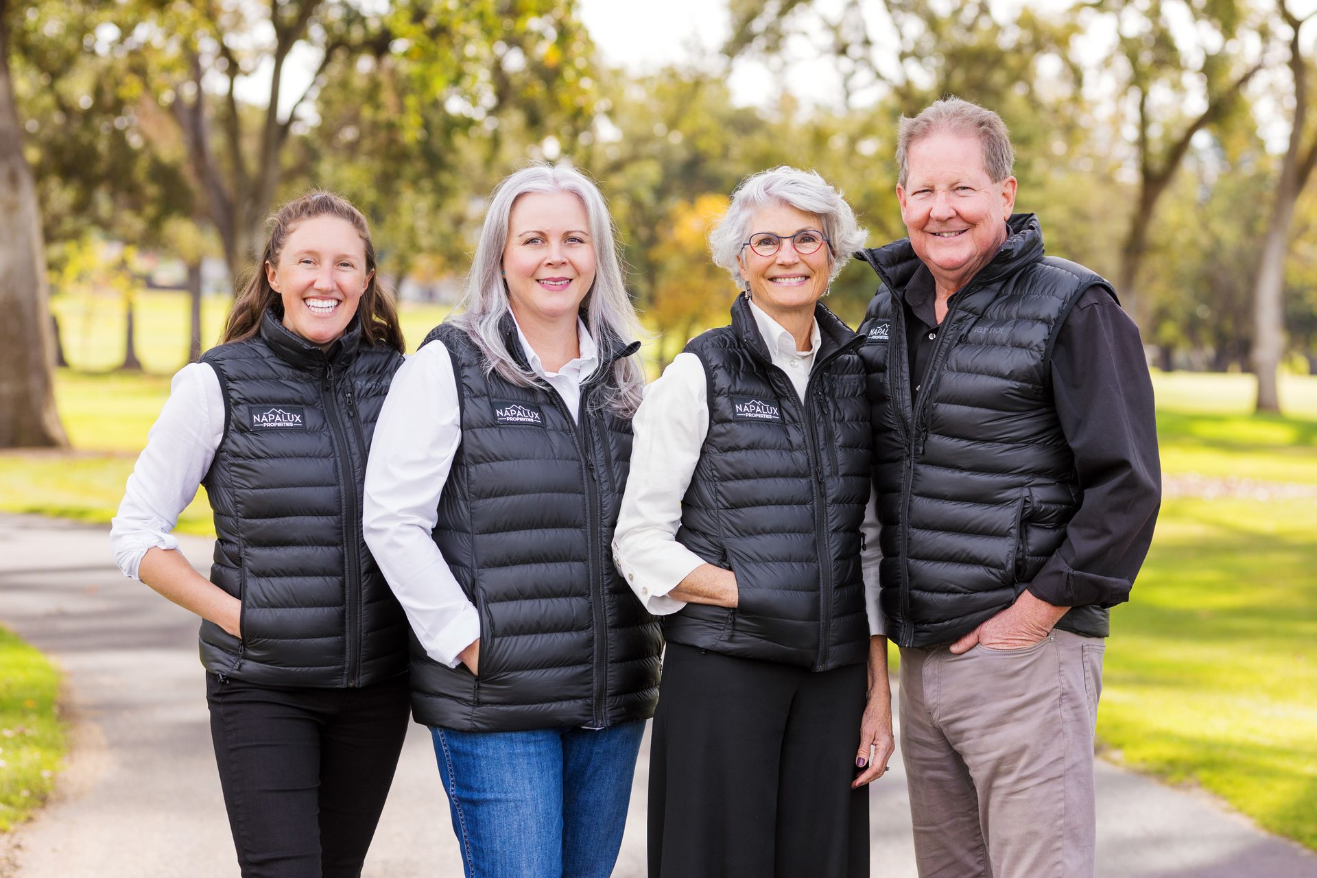 Four people standing outdoors on a tree-lined path, wearing black vests and smiling at the camera.