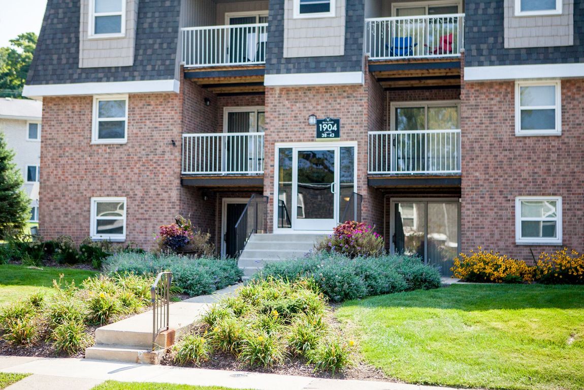 Apartment building with brick facade, multiple levels, balconies, and entrance stairs. Green bushes and grass surround.