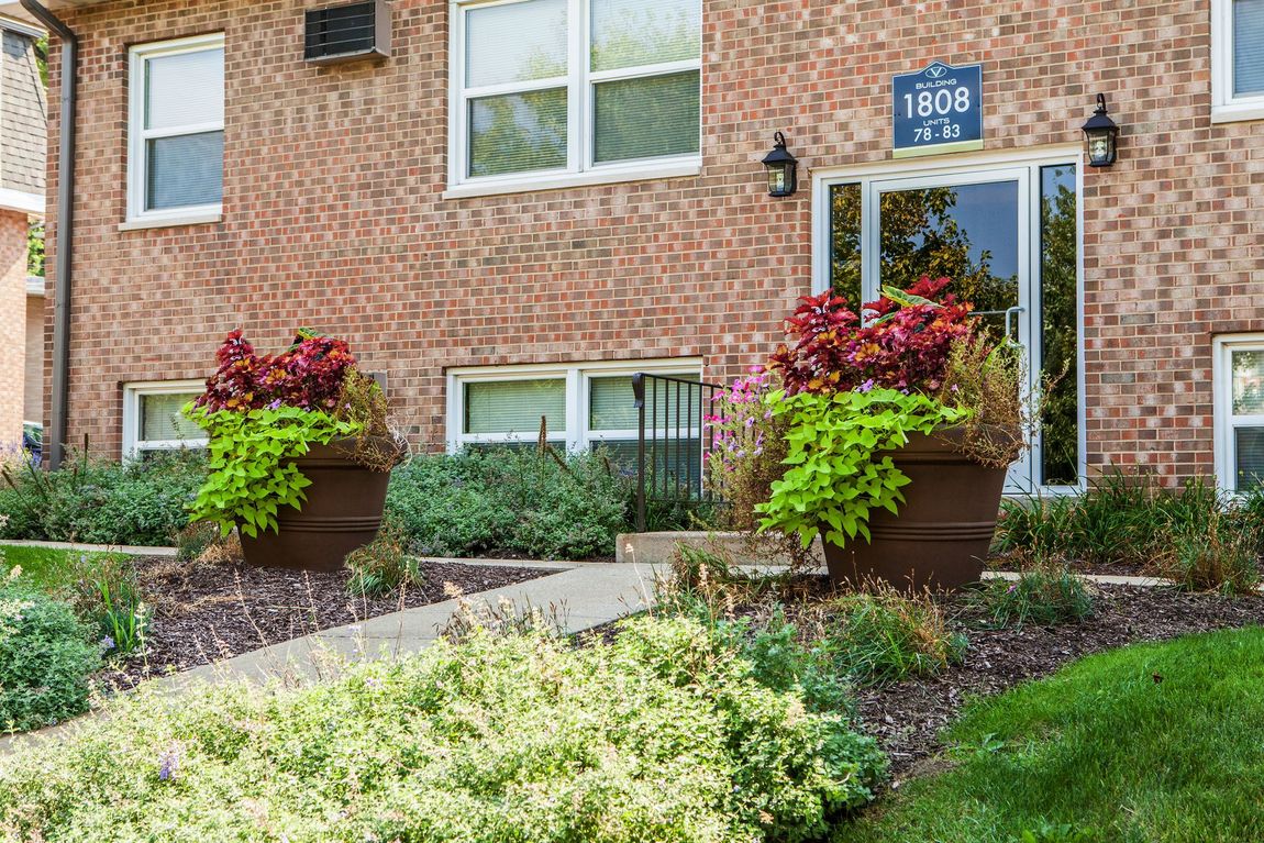 Apartment building exterior with brick facade and landscaped entrance; planters, pathway, and windows visible.