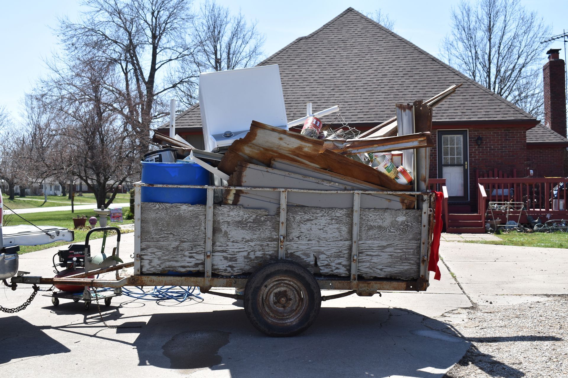 A utility trailer packed with debris and a blue plastic barrel, parked on a paved driveway in front of a brick house. A utility trailer packed with debris and a blue plastic barrel, parked on a paved driveway in front of a brick house.