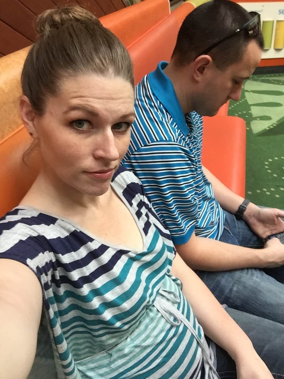 Two people seated on orange benches beside green water, one taking a selfie and the other looking at a phone.