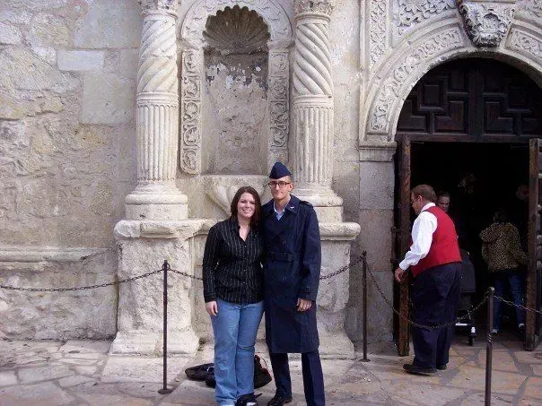 Two people posing in front of an ornate stone building entrance, with a man standing near the doorway.