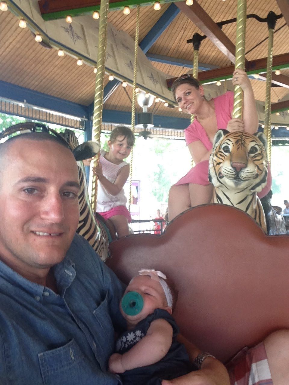 Man holding a baby on a carousel tiger with two people smiling in the background
