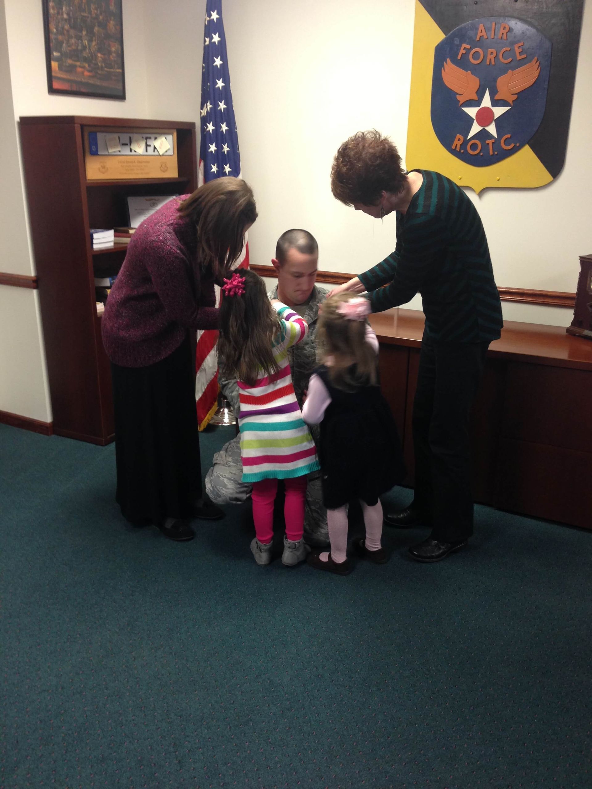 Four people stand in an office, looking at a child near a counter beneath a U.S. Air Force emblem.