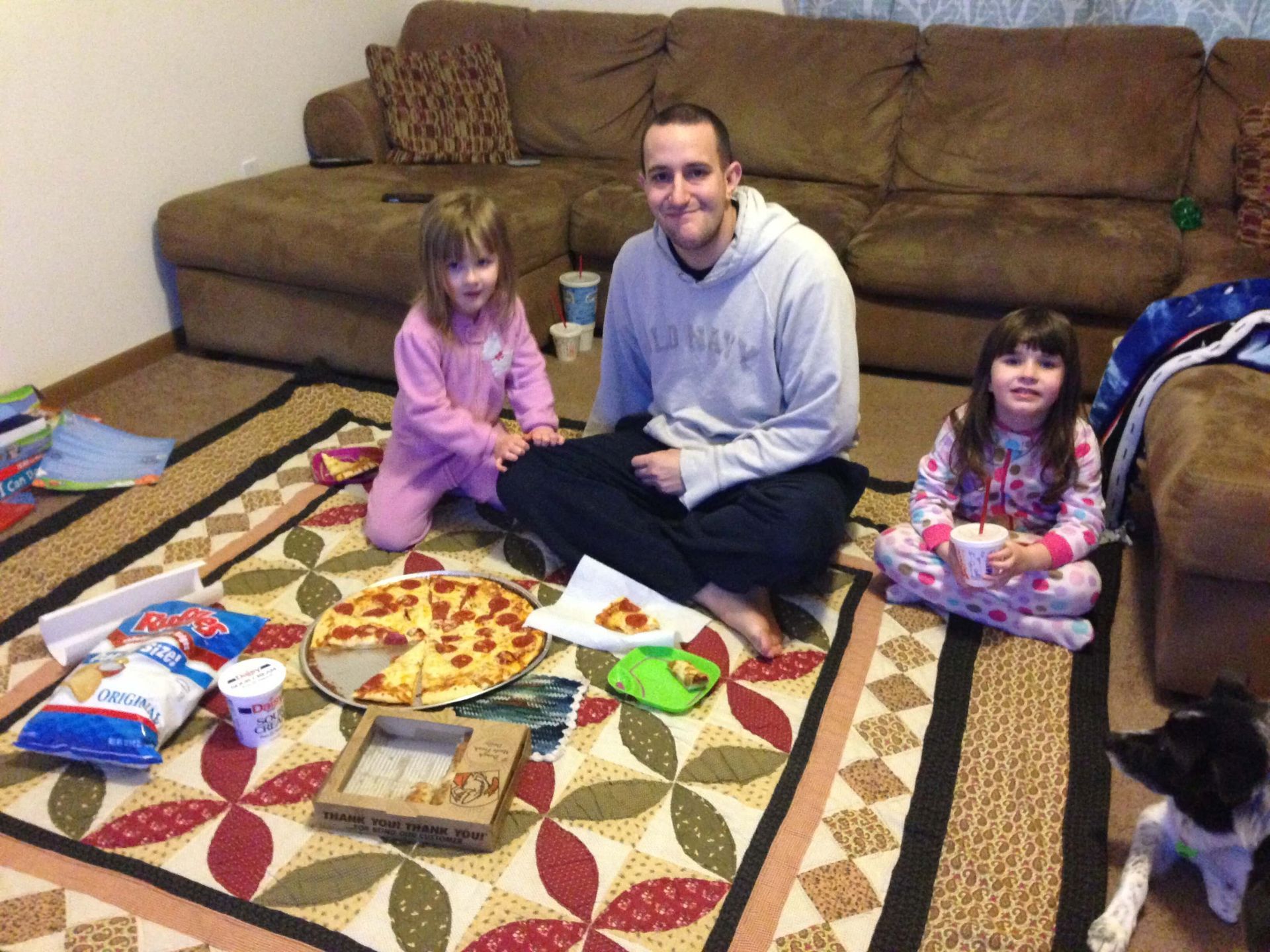 Family sitting on a living room rug with pizza and snacks, smiling near a brown couch