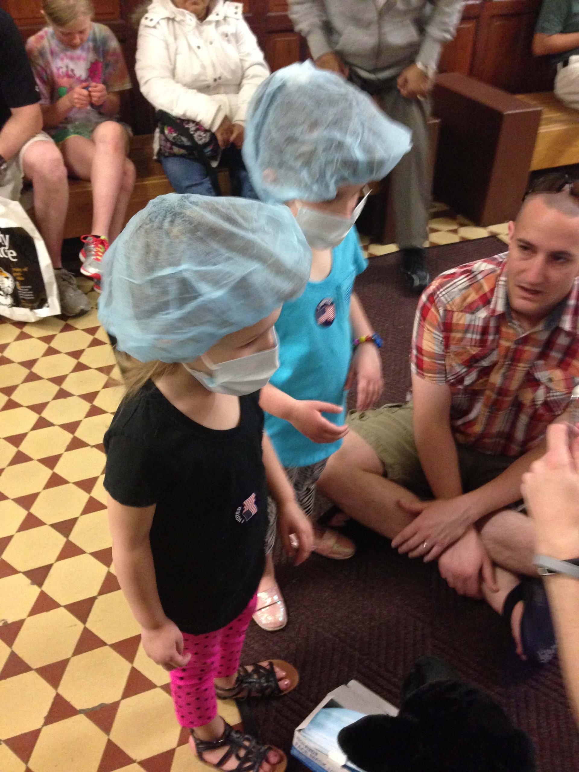 Two children in blue hairnets stand with a seated man in a tiled indoor room.