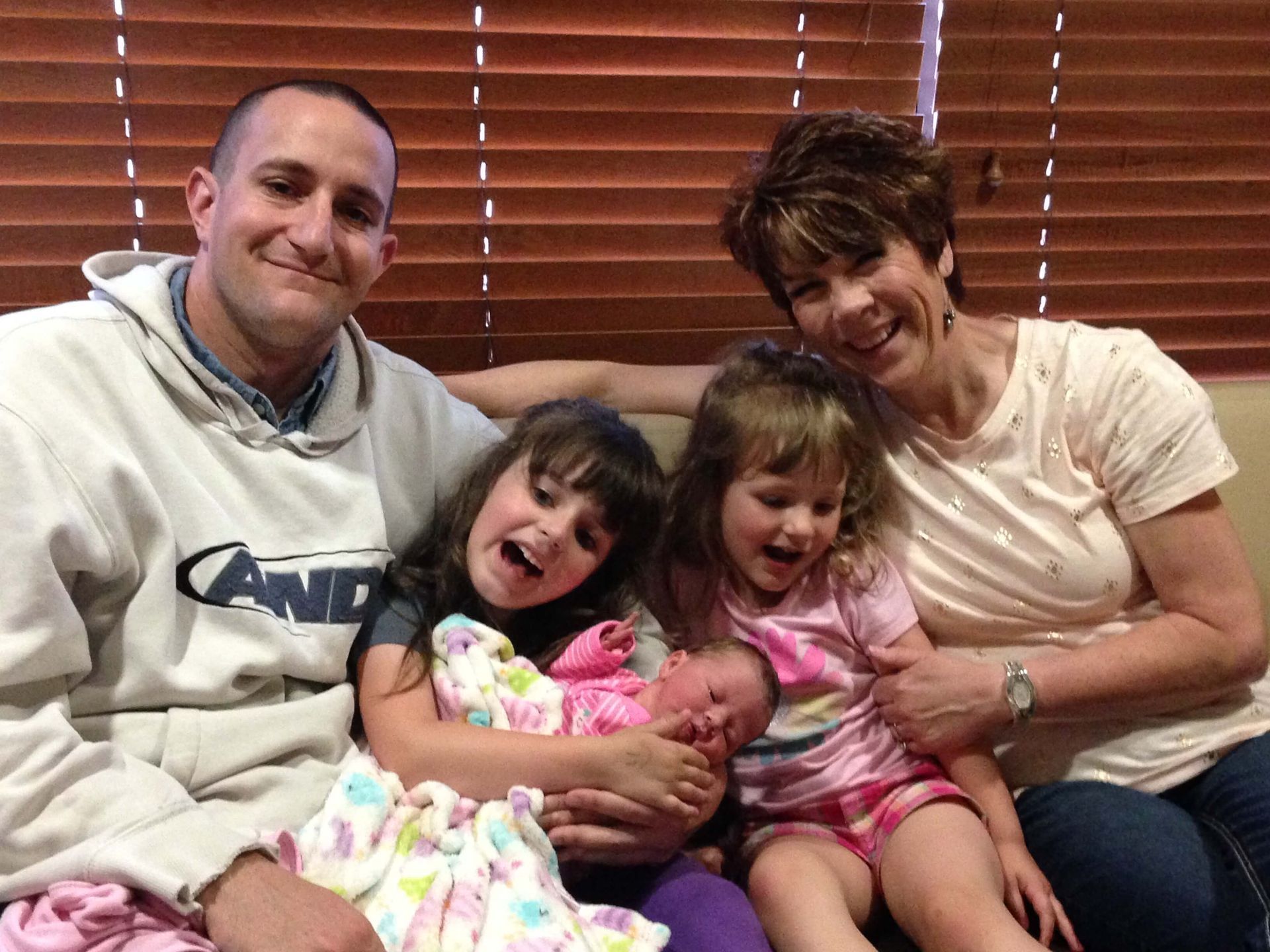 Family sitting on a couch, smiling and holding a newborn baby indoors by the window.
