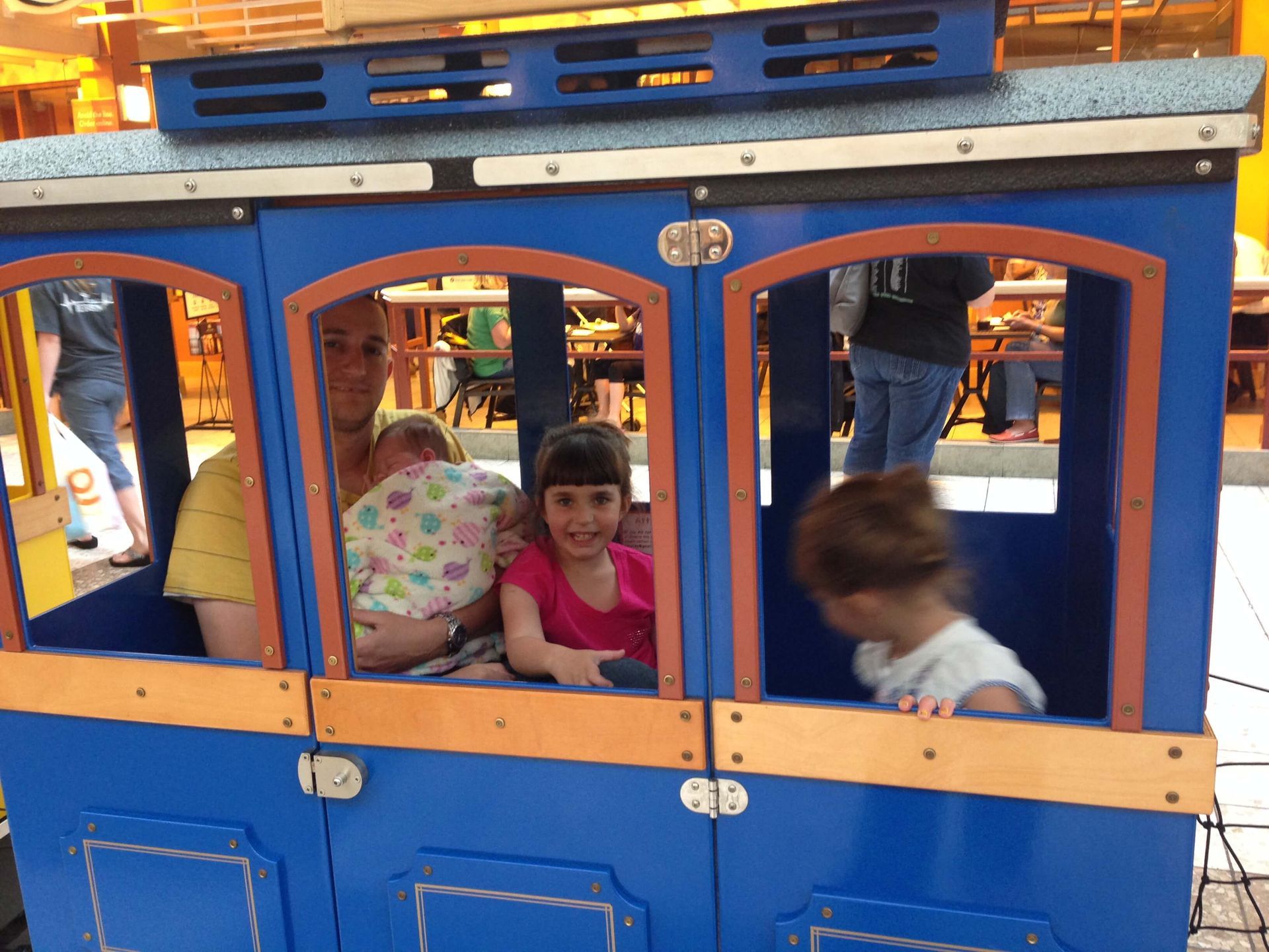 Children riding in a blue-and-orange play train at an amusement park, smiling inside the car.