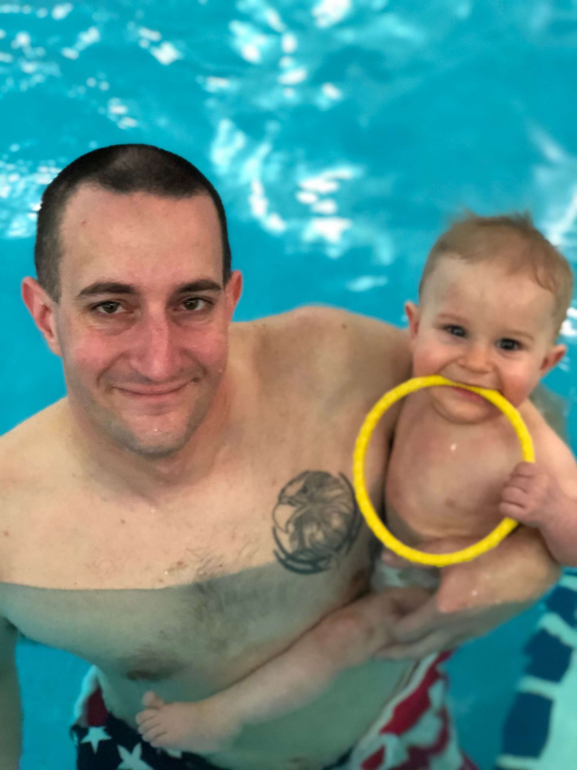 Two people in a swimming pool, one holding a baby with a yellow ring toy around the baby’s arm.
