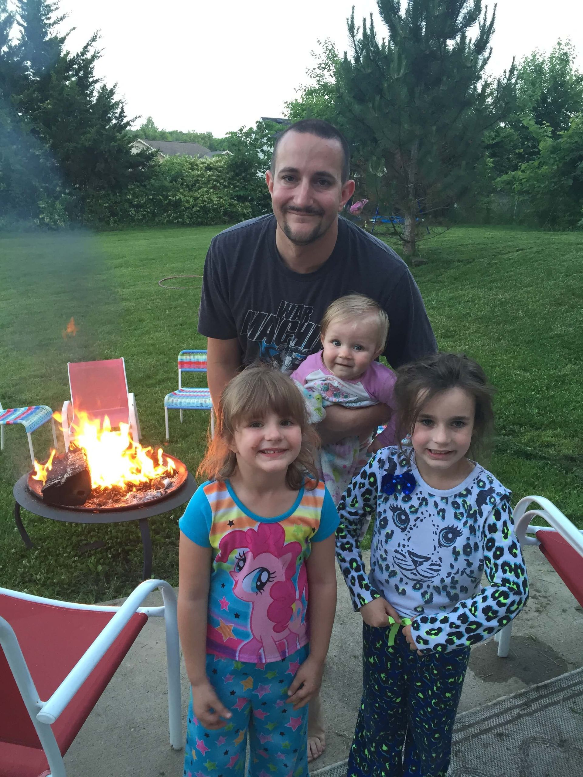 Three children and an adult smiling outdoors by a glowing fire pit in a backyard.