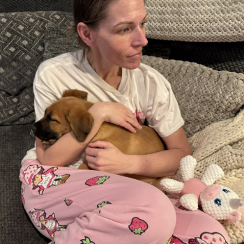 Woman cuddles a puppy on a couch beside a crocheted stuffed toy, wearing pink pajama pants.