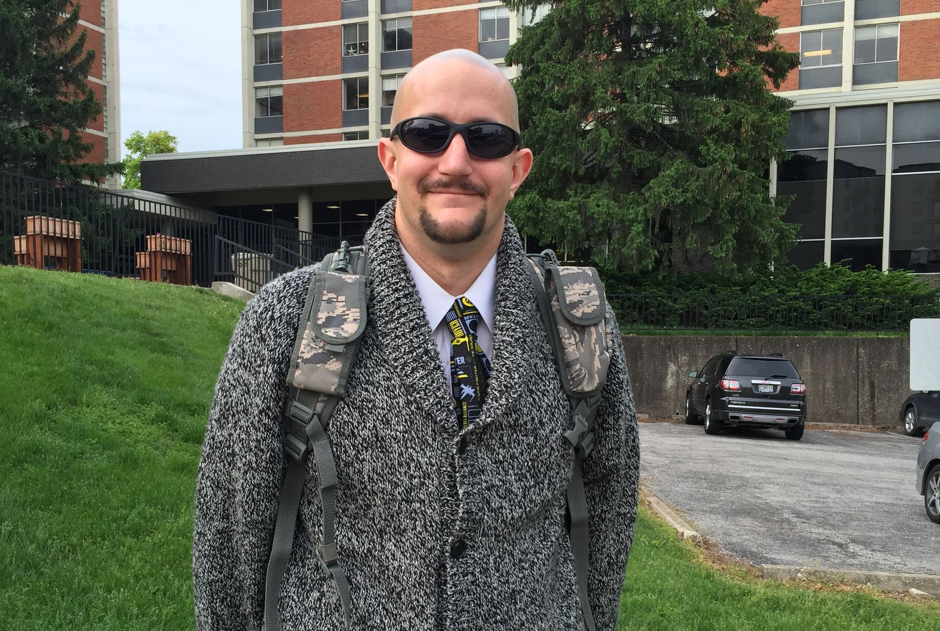 Smiling man in sunglasses and a gray coat standing outdoors near a brick building and trees.