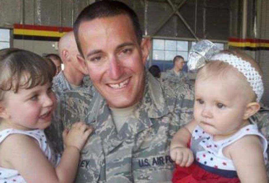 Smiling U.S. Air Force servicemember holding two young children indoors at a hangar.