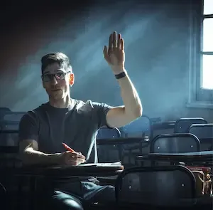Person seated in a dim classroom, raising a hand at a desk by the window.