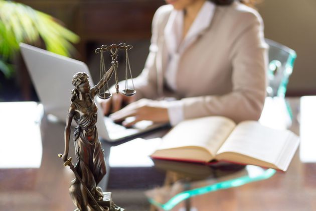 Three people in business attire at a table: Two facing each other, and one in the background holding papers. Three people in business attire at a table: Two facing each other, and one in the background holding papers.