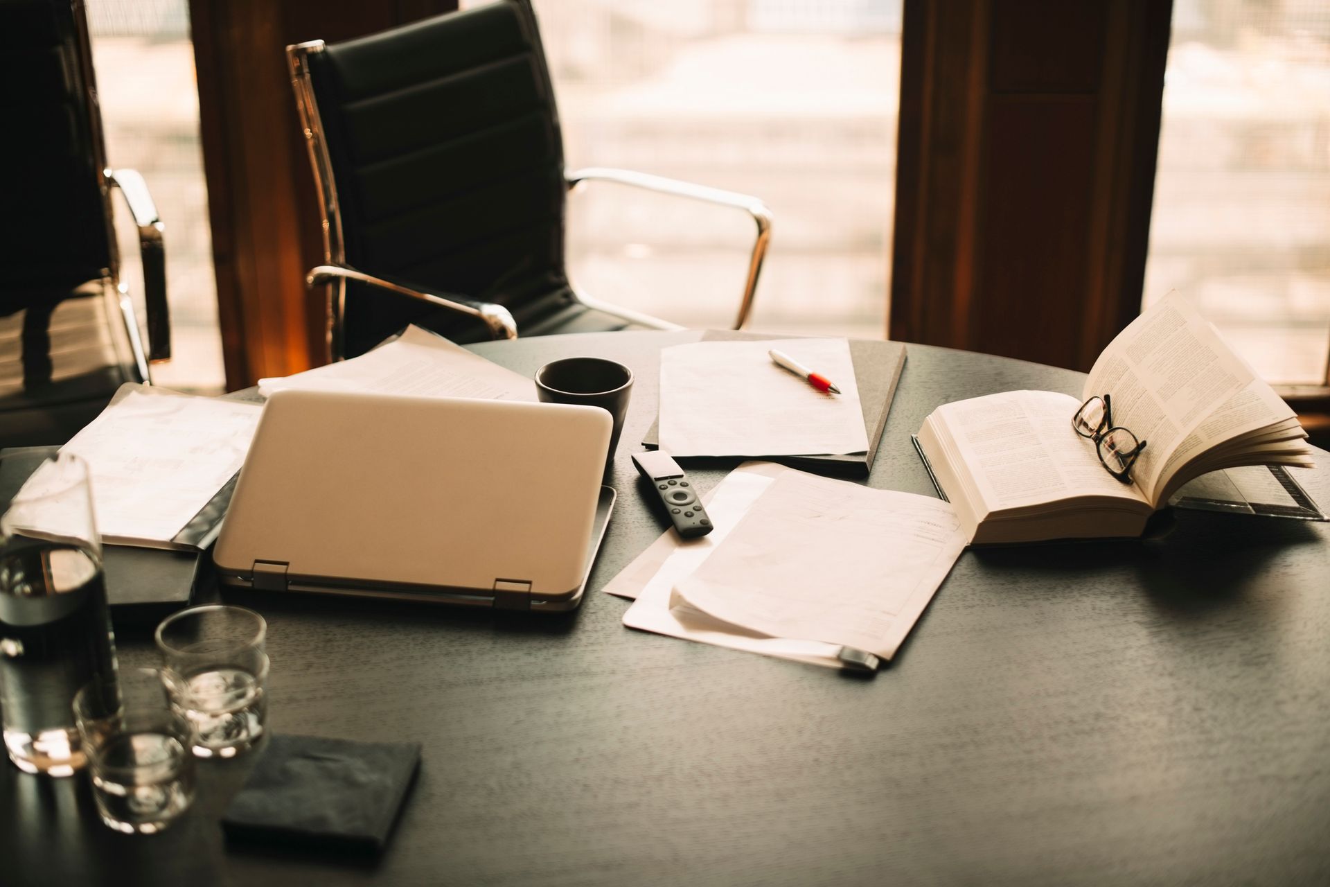Desk with laptop, papers, book, glasses, water glasses, and a chair near a window.