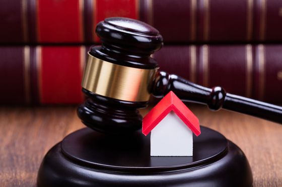 Gavel striking a small house figurine on a wooden block, with law books in background.
