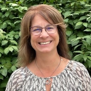 Woman with glasses smiling in front of green foliage. She wears a patterned blouse and necklace.