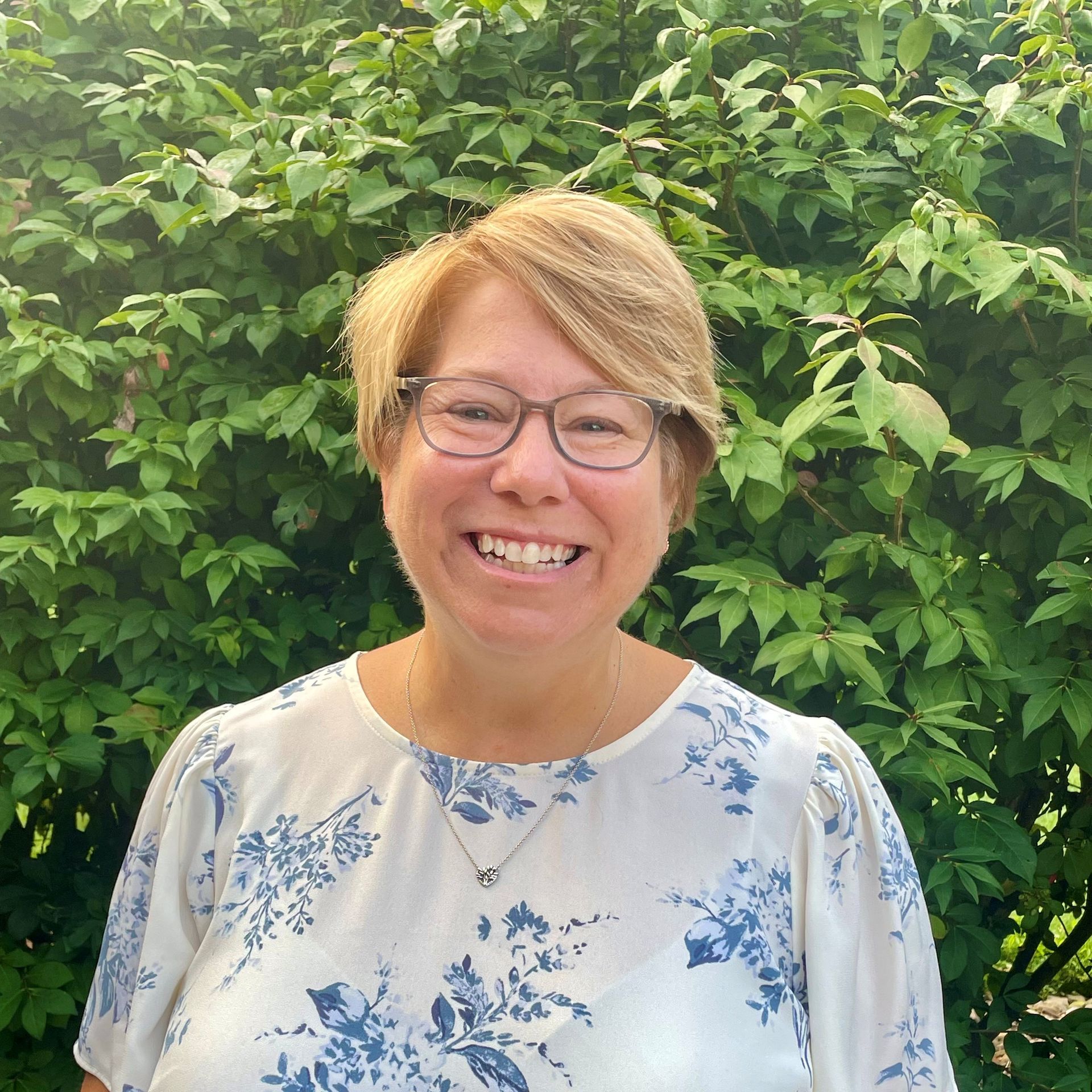 Woman with short blonde hair and glasses smiles in front of green foliage, wearing a floral blouse.